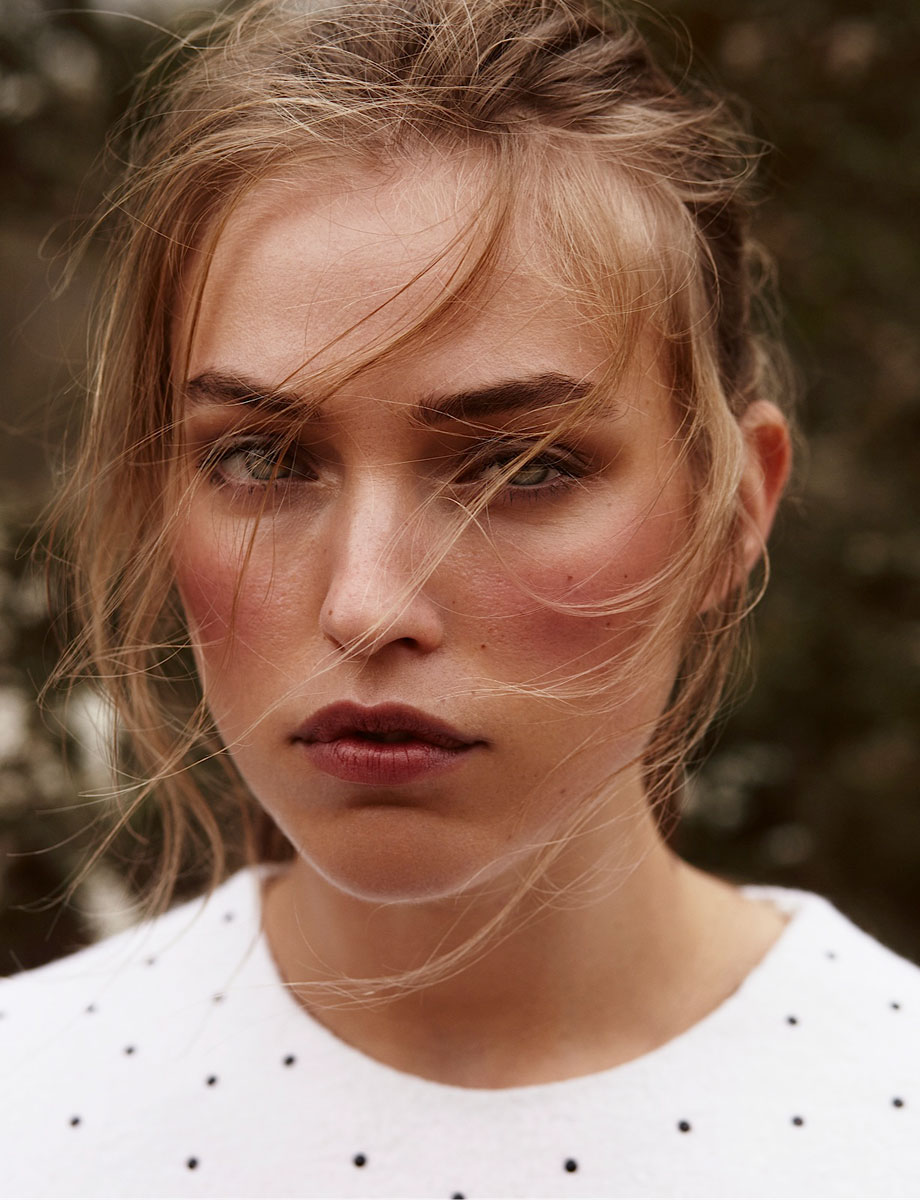 Close-up portrait of a young adult woman with blond hair, wearing a dotted shirt, set against a blurred outdoor background.