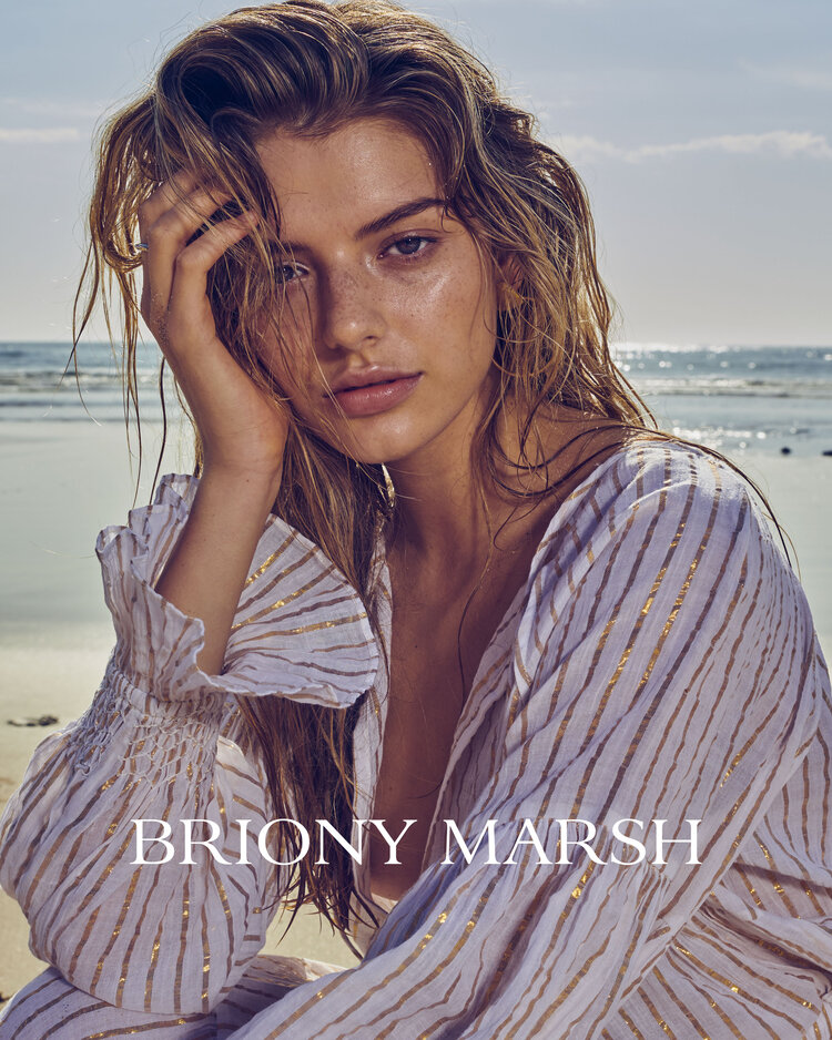 A woman with wet blonde hair and a white blouse with gold stripes poses against a beach and ocean backdrop on a sunny day.