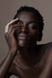 Close-up portrait of a Black woman with short dark hair, hand raised to her face with metallic gold paint on her fingers, against a neutral studio backdrop.