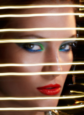Close-up of a woman's face with bold makeup, partially obscured by horizontal gold lines casting shadows and creating a blurred effect.