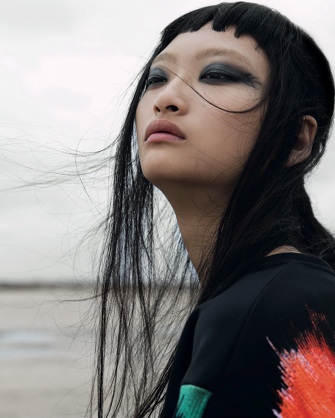 Asian woman gazes up in black top with red accents against a neutral background. Her dark hair blows in wind and heavy dark eye makeup.