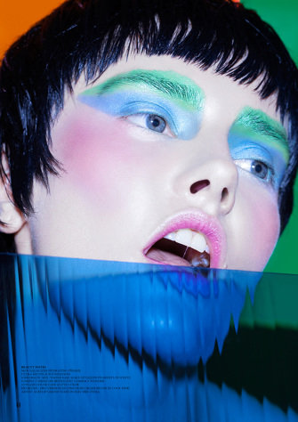 Close-up portrait of a woman with colorful makeup and short black hair, wearing blue plastic, with a blurred green and orange background.
