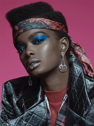 Headshot of a woman with blue eye shadow and silver lips wearing a patterned headscarf, statement earrings, and a black quilted jacket.