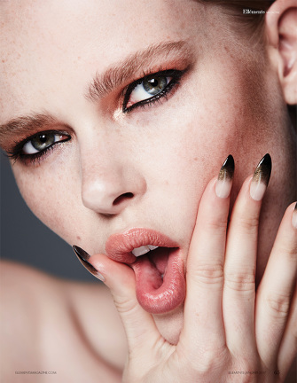 Close-up portrait of a young woman with creative eyeshadow, long nails, and her tongue sticking out, shot indoors.