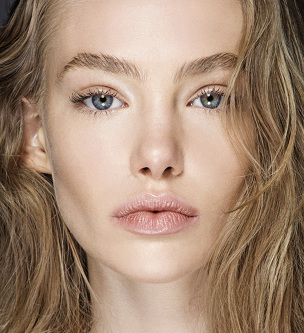 Close-up portrait of a blonde woman with blue eyes and light pink lips, emphasizing skincare and natural beauty in a studio setting.