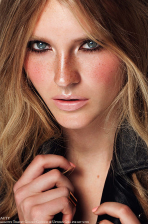 Close-up portrait of an adult female with light brown wavy hair, freckles, makeup, and a black jacket. Beauty and fashion portrait.