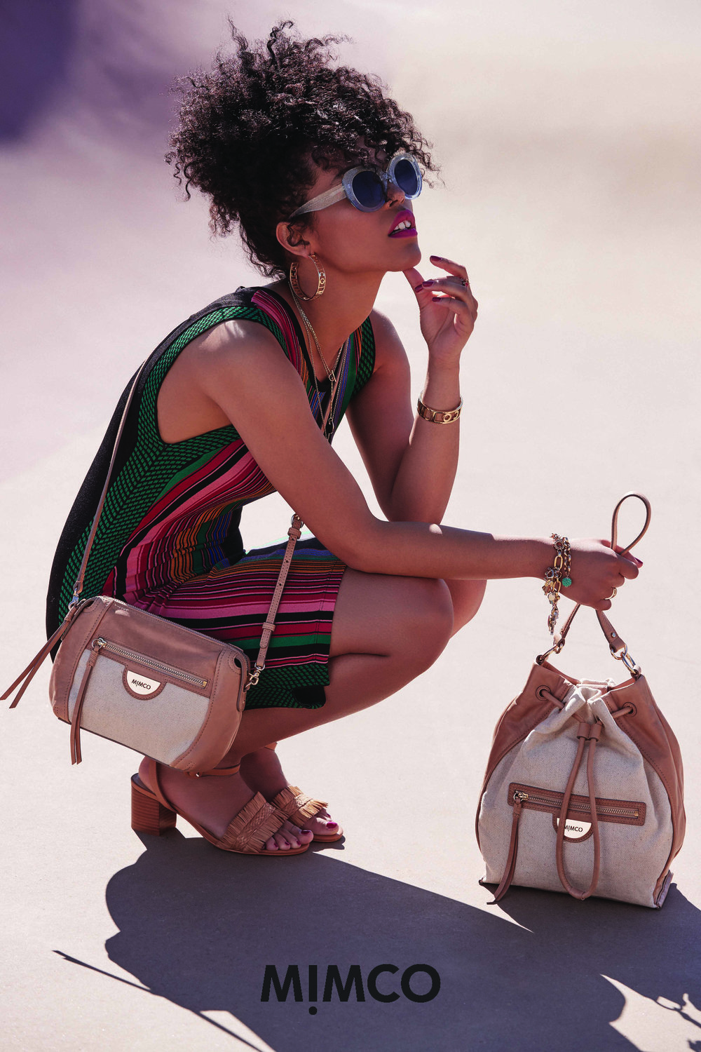 Woman models Mimco purse and handbag while crouching outside in sunglasses and a striped dress, accessorized with jewelry.