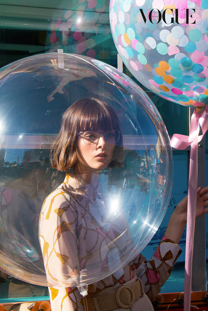 A woman in a printed dress stands inside a clear bubble with a large confetti balloon on a sunny day, a vogue magazine logo is visible.