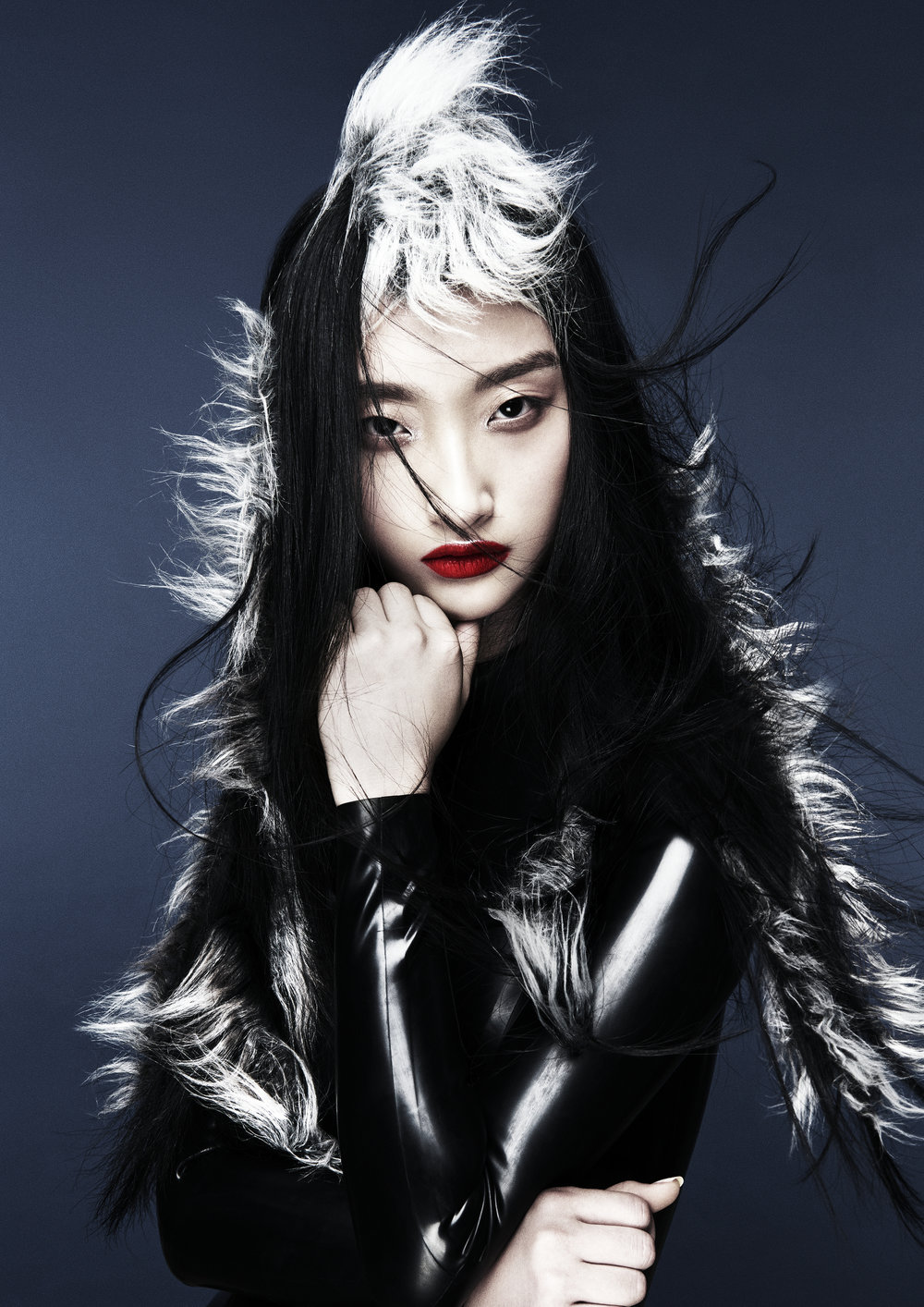 Close-up portrait of a stylish Asian woman with black and white hair, red lipstick, and a black faux fur coat, against a gray backdrop.