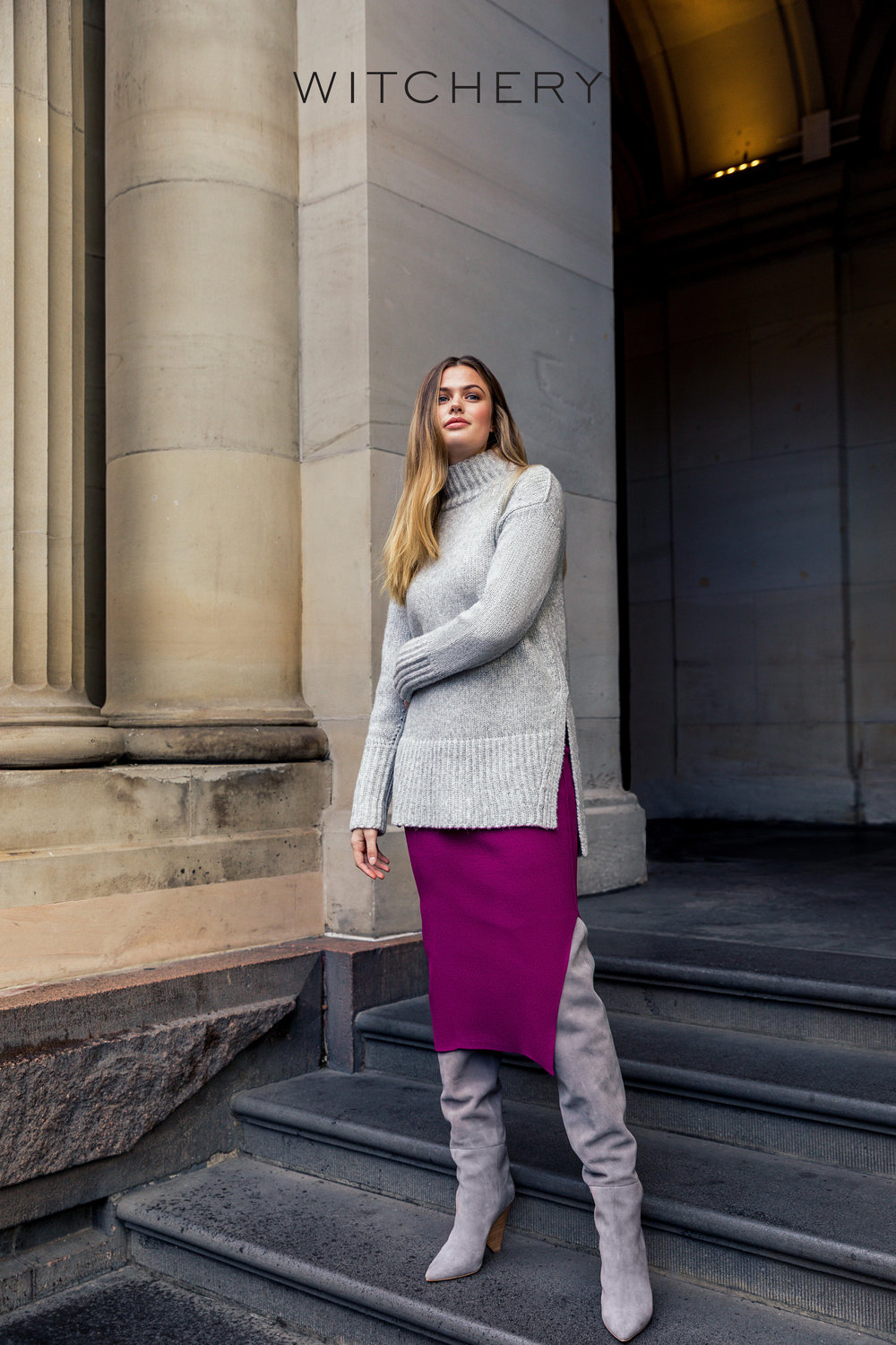 Adult female poses on stone steps wearing a grey turtleneck sweater, magenta skirt, and grey thigh-high boots near a stone column.