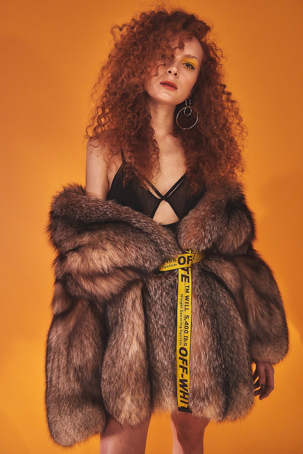 A young adult woman with curly red hair poses with a serious expression wearing a fur coat over a black top against an orange background.