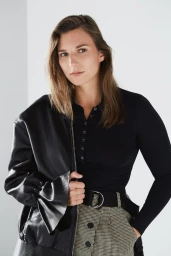 Portrait of a woman wearing a black top and patterned trousers with a black jacket draped over her shoulder against a white background