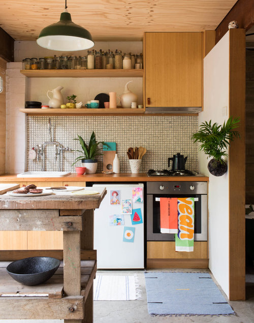 Rustic modern kitchen interior with wooden cabinets, vintage table, plants, green pendant light, tiled backsplash, and various kitchenware.