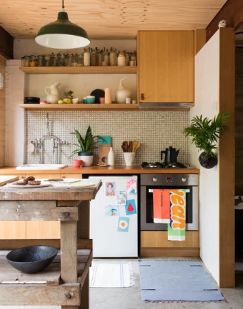 Rustic modern kitchen interior with wooden cabinets, vintage table, plants, green pendant light, tiled backsplash, and various kitchenware.