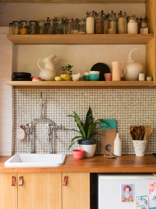 Bright, airy kitchen featuring wooden shelves stocked with jars, white ceramic pitchers and mugs, a potted plant, and a white sink.