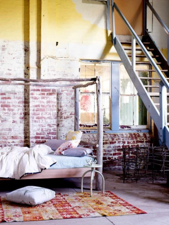 Rustic bedroom interior with brick walls, distressed plaster, four-poster bed, patterned rug, natural light, and stairway.