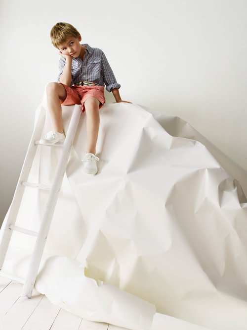 Young boy with blond hair in button-down shirt and shorts sits on a ladder in a studio with a large white backdrop.