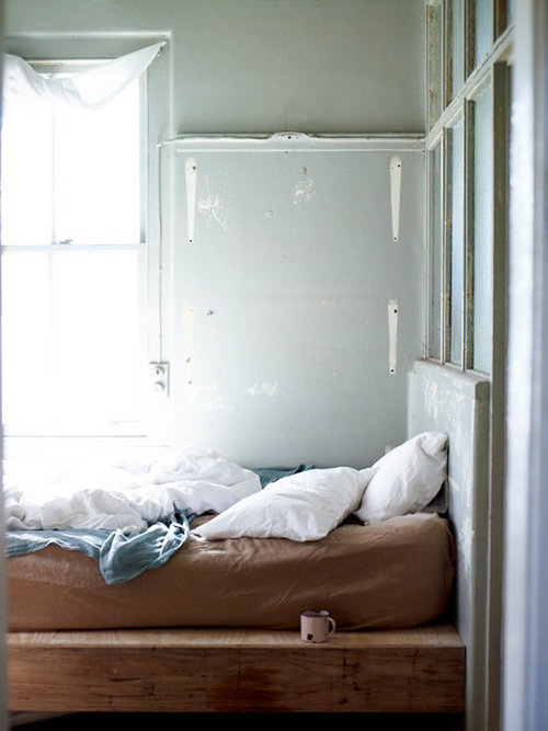 Bedroom interior featuring a bed with white linens, wooden frame, pale green walls, and natural light from a nearby window. Cozy and inviting.