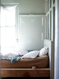 Cozy bedroom interior styled with a wooden platform bed, white pillows, brown duvet, and soft natural window light.