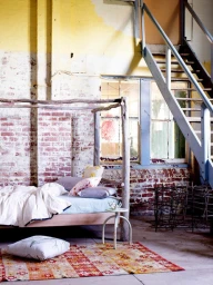 Rustic wooden canopy bed styled with patterned pillows in an industrial loft interior with exposed brick and stairs