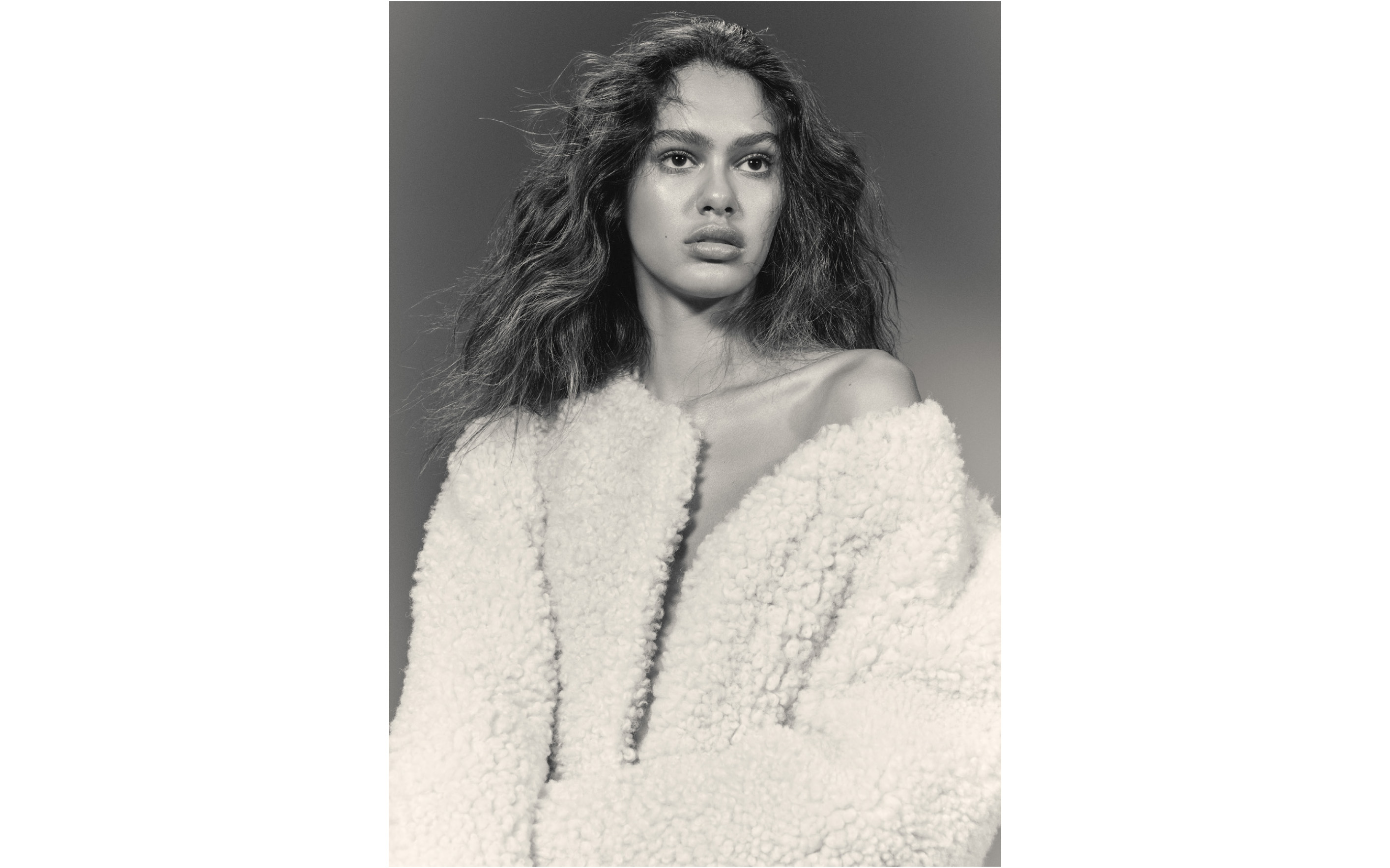 A young adult woman poses indoors in a fluffy, oversized off-white jacket, with natural makeup and wavy hair against a plain grey background.