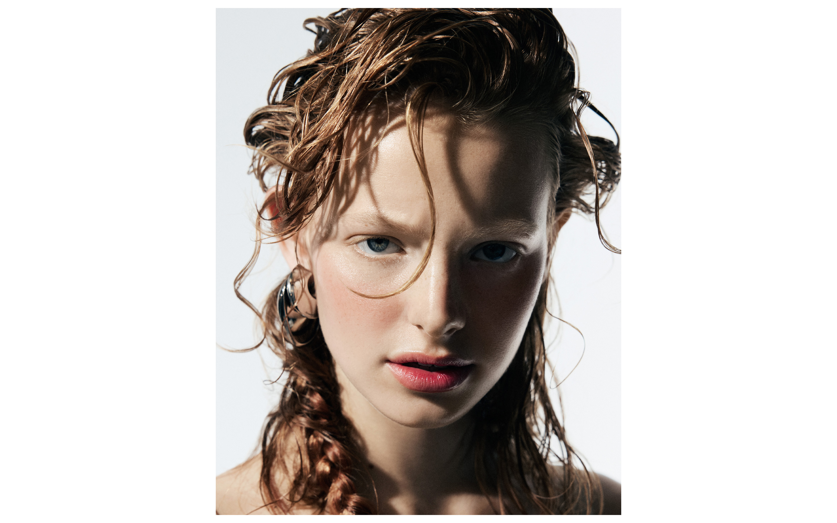 Close-up portrait of a woman with wet, styled hair, light skin, and soft makeup against a bright, neutral backdrop in a beauty editorial.