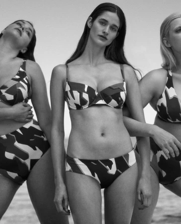 Three female models wearing patterned two-piece swimwear against an outdoor sky background in a black and white photograph.