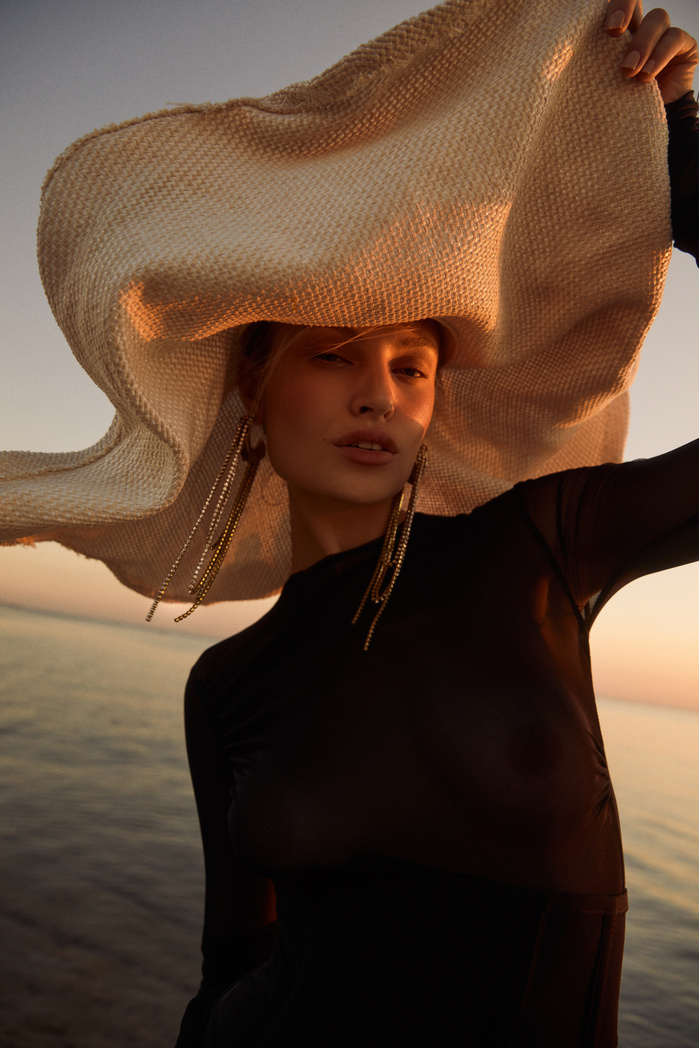 A woman poses at sunset wearing a large sun hat and dark dress, complemented by long earrings, with a soft ocean background.
