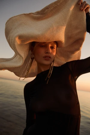 Woman in sheer black top wearing an oversized straw sun hat holding the brim against a sunset sky and ocean background.
