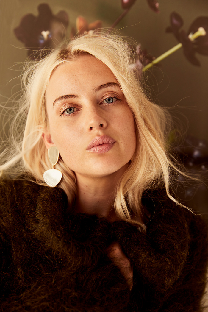 Portrait of a woman with blonde hair wearing gold earrings and a green sweater in front of a backdrop with dark flowers.