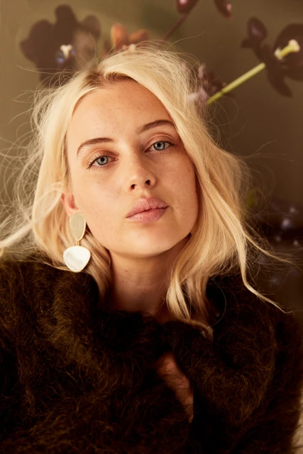 Portrait of a woman with blonde hair wearing gold earrings and a green sweater in front of a backdrop with dark flowers.