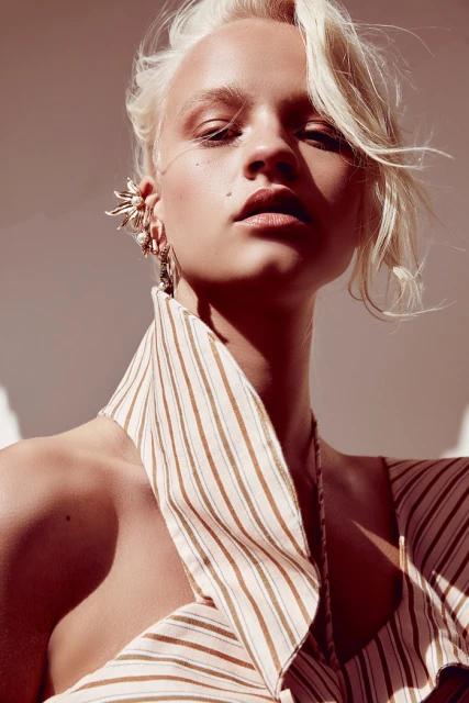 Close-up fashion portrait of an adult female with short blonde hair wearing an ear cuff and a striped halter top against a plain backdrop.