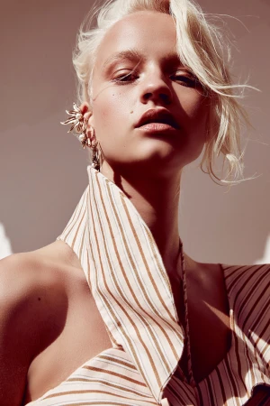 Close-up fashion portrait of an adult female with short blonde hair wearing an ear cuff and a striped halter top against a plain backdrop.