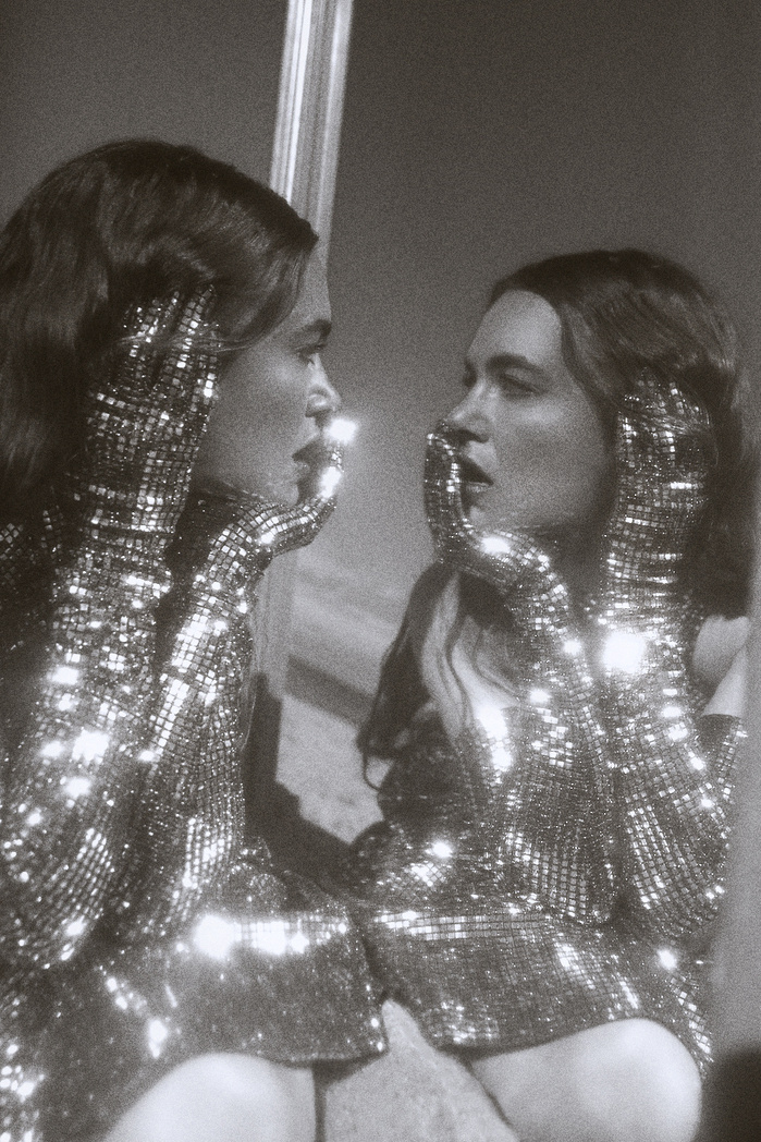 A woman in a sparkling dress and gloves looks into a mirror, reflecting her image in a black and white, vintage-style photograph.