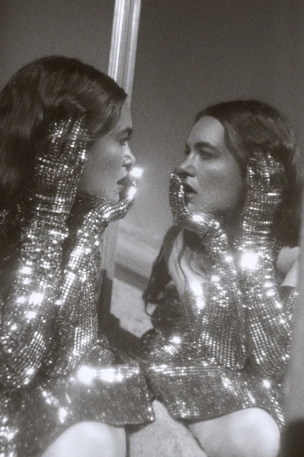 A woman in a sparkling dress and gloves looks into a mirror, reflecting her image in a black and white, vintage-style photograph.