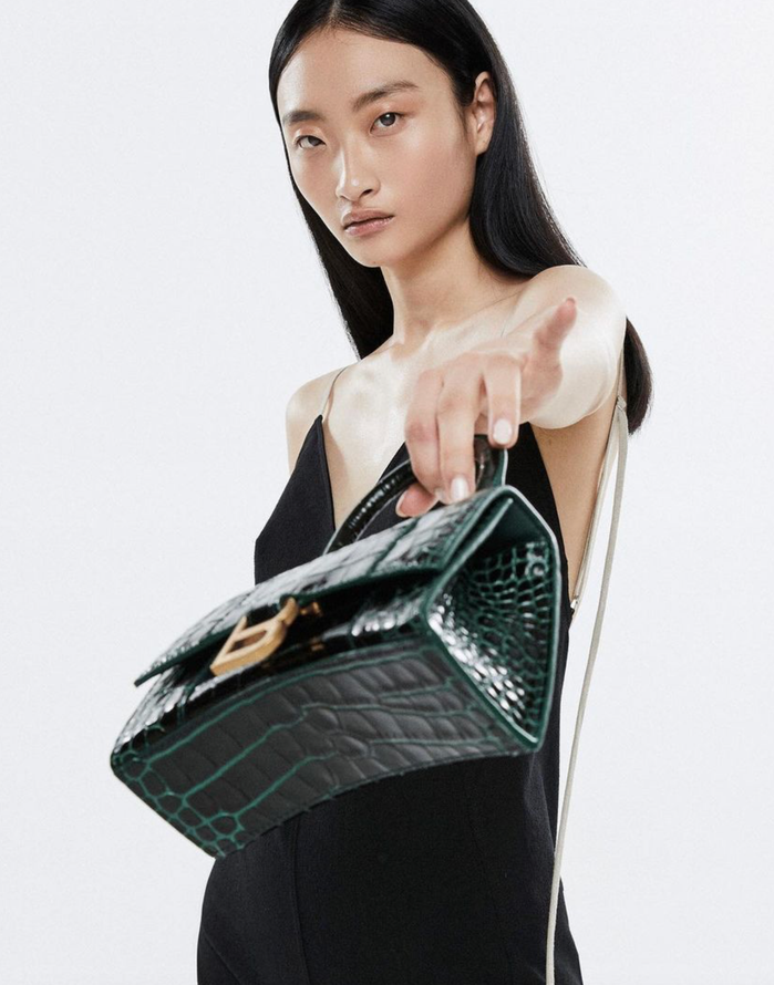 A woman models a green crocodile-textured Balenciaga purse, wearing a black dress against a white backdrop in a fashion setting.