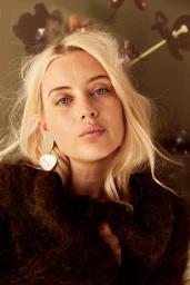 Close-up portrait of a blonde woman wearing statement drop earrings and a textured dark garment.