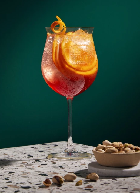 Cocktail in a stemmed glass with orange slices and a twist, garnished with pistachios, on a terrazzo table against a green background.