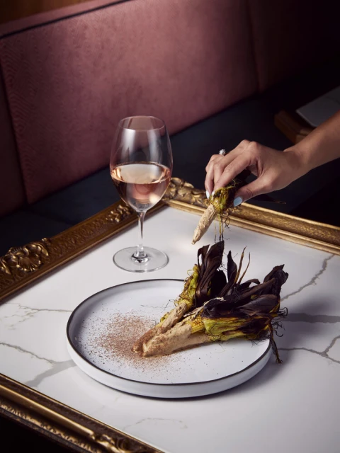 Elevated food presentation showcasing two arranged corn cobs dusted with brown powder, wine glass, and a hand adjusting one of the cobs.