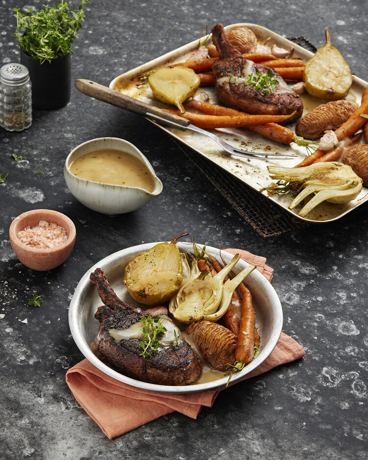 Overhead shot of cooked pork chop with roasted vegetables and pears on plates and trays, presented on a dark stone surface.