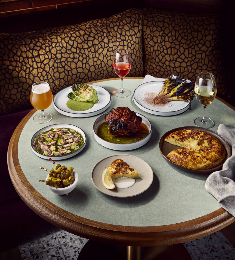 Overhead shot of round table with several plates of diverse dishes, assorted glassware, brown and tan patterned bench in background.