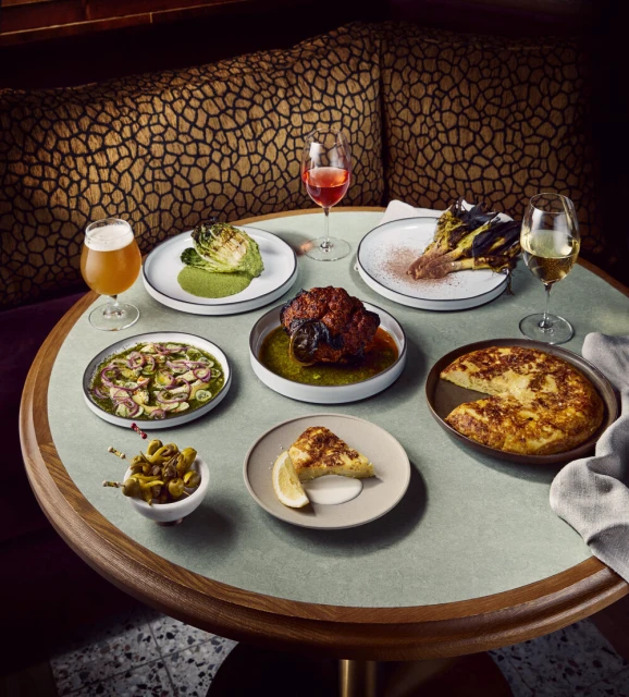 Overhead shot of round table with several plates of diverse dishes, assorted glassware, brown and tan patterned bench in background.