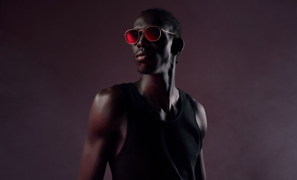 Portrait of an adult Black male wearing a black tank top and sunglasses with a red tint on a dark background in a photo studio.