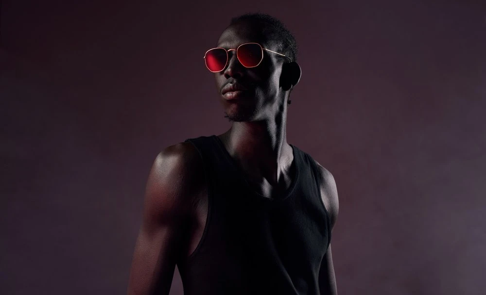 Portrait of an adult Black male wearing a black tank top and sunglasses with a red tint on a dark background in a photo studio.