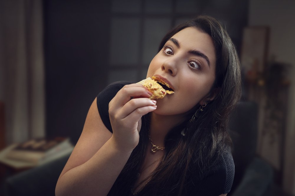 Close-up of a woman with wide eyes eating food indoors, wearing a black shirt and gold jewelry, with blurred background details.