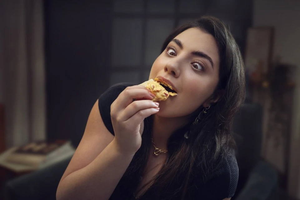 Close-up of a woman with wide eyes eating food indoors, wearing a black shirt and gold jewelry, with blurred background details.