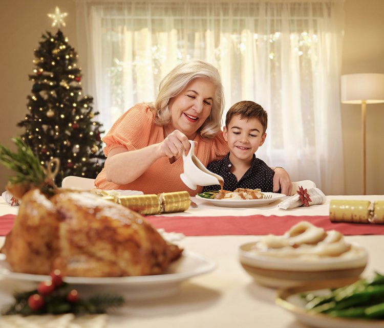 Grandmother and grandson share holiday meal together at a decorated table with a Christmas tree in the background at home.