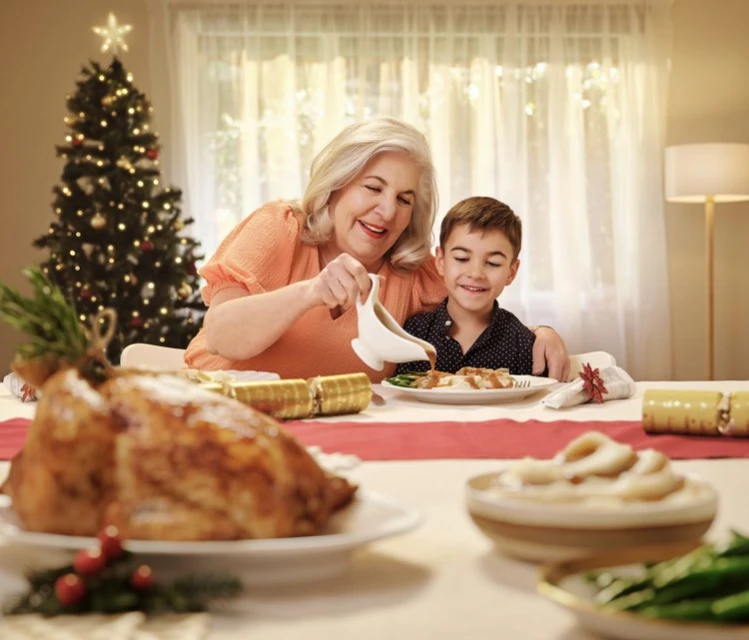 Grandmother and grandson share holiday meal together at a decorated table with a Christmas tree in the background at home.