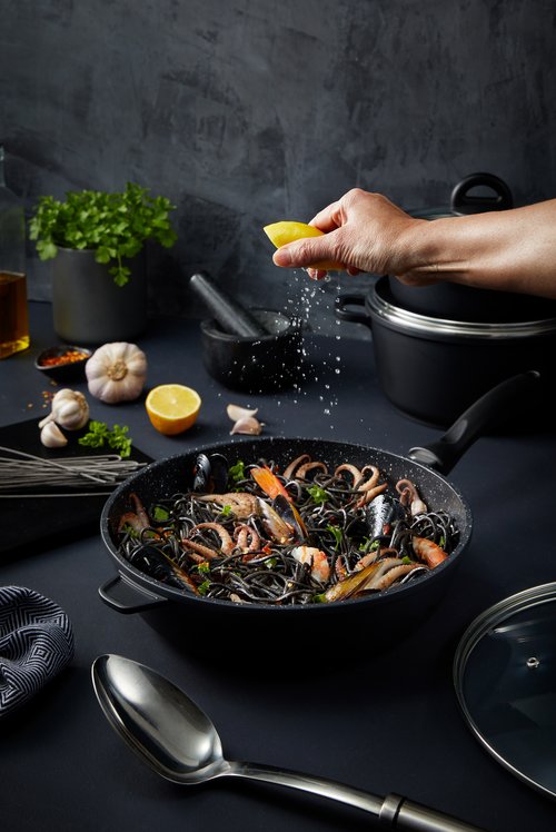 Overhead shot of black spaghetti with seafood in a pan being seasoned with lemon, surrounded by cooking utensils and ingredients.