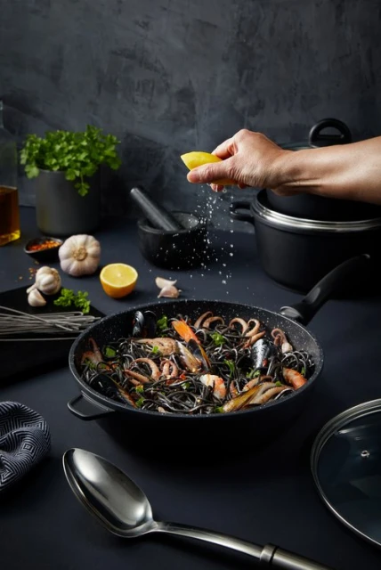 Overhead shot of black spaghetti with seafood in a pan being seasoned with lemon, surrounded by cooking utensils and ingredients.